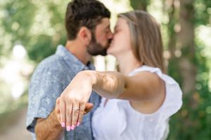A couple shares a romantic engagement moment while showing off a ring in a sunlit outdoor setting.