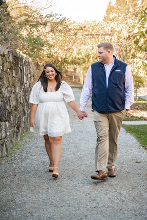 A couple walks hand in hand along a stone wall path during their engagement session.