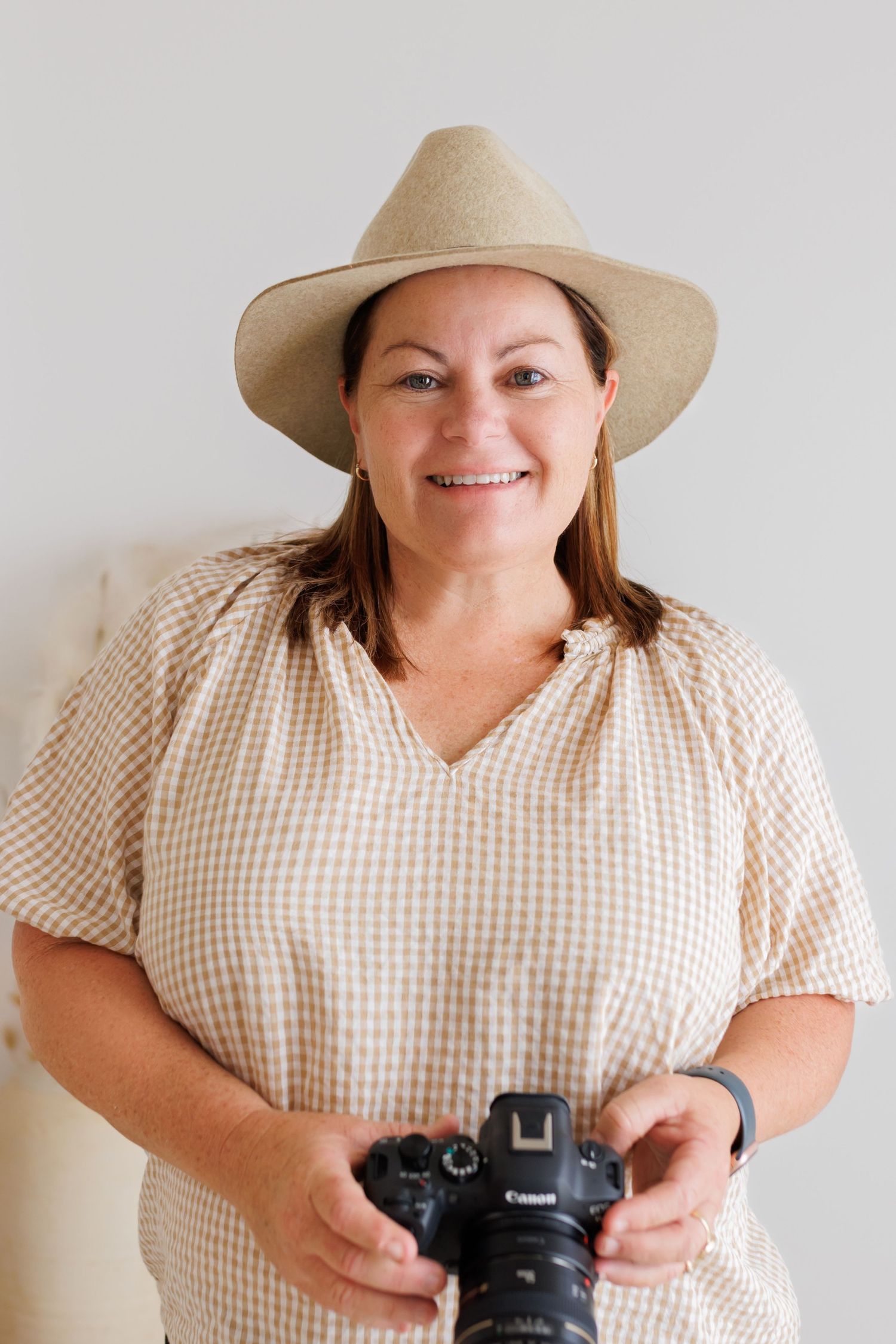 Photographer in gingham dress and straw hat smiling while holding camera against white background.