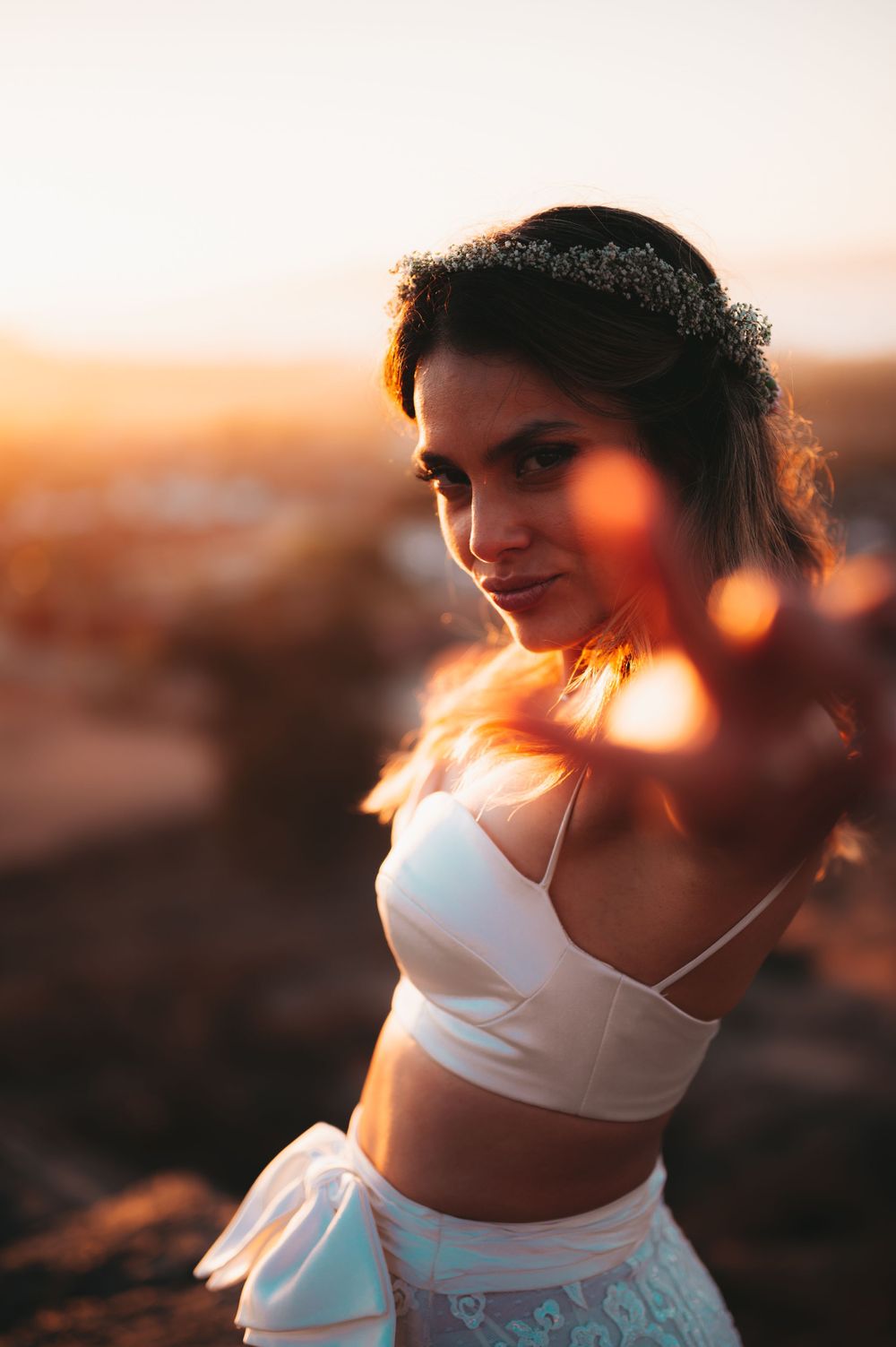 Bride in white crop top and skirt wearing floral crown poses at golden sunset with warm lens flare.