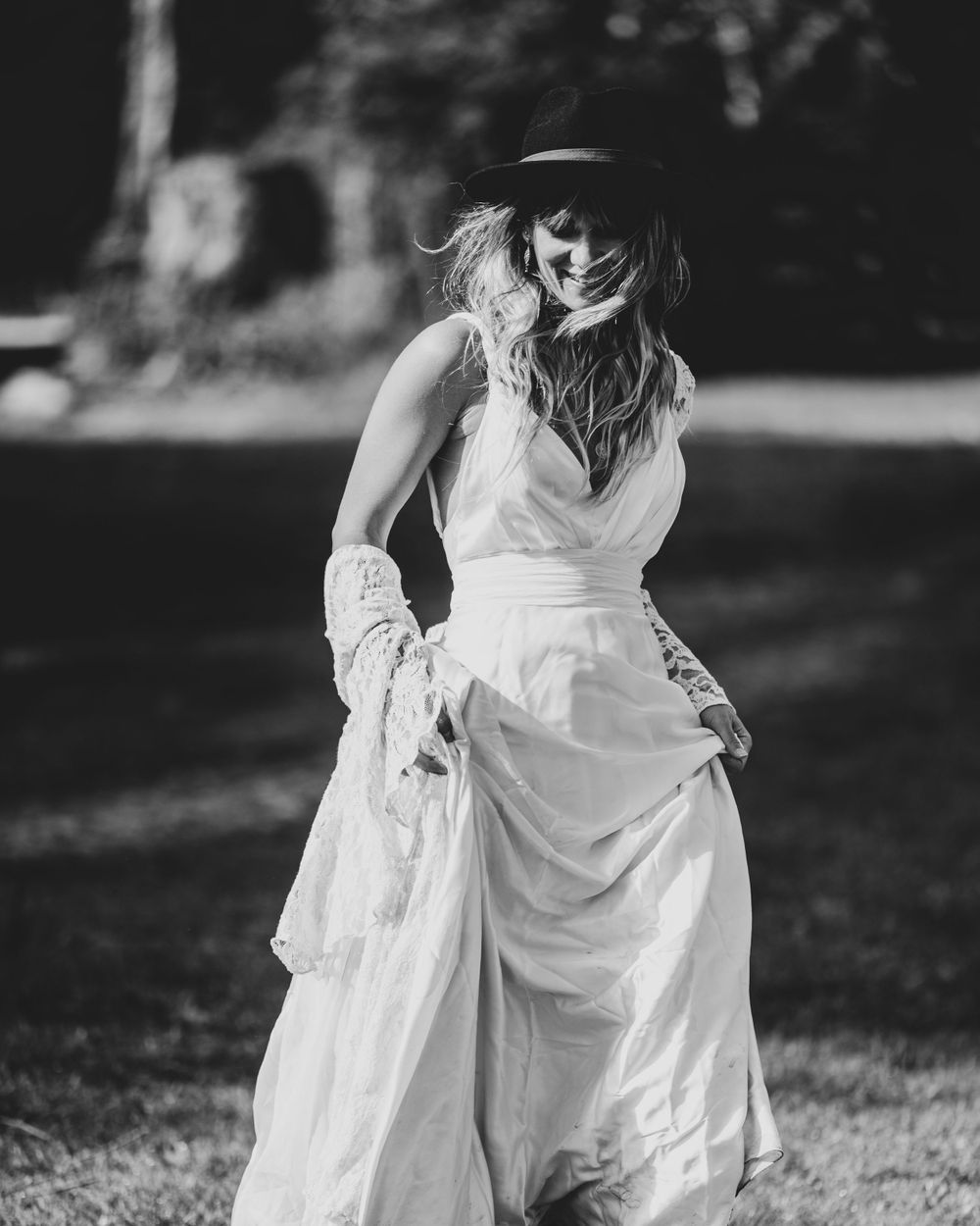 Black and white photo of a smiling bride in a white dress and black hat outdoors.