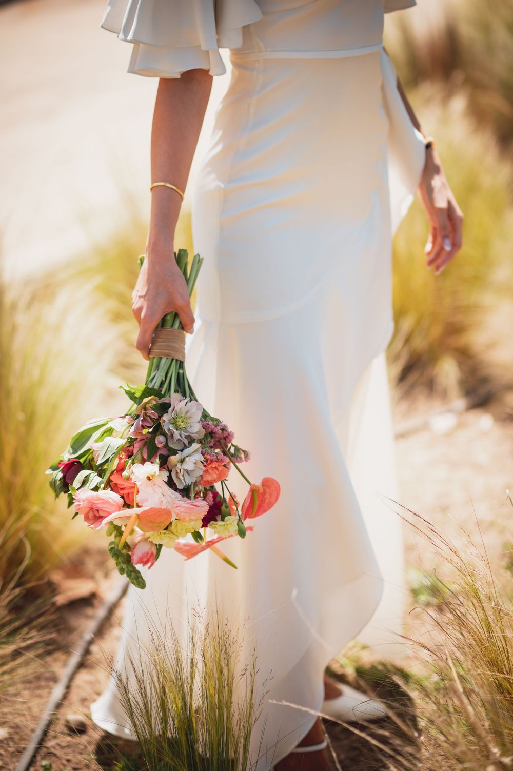 Bride in white dress holding colorful bouquet of pink and coral flowers outdoors.