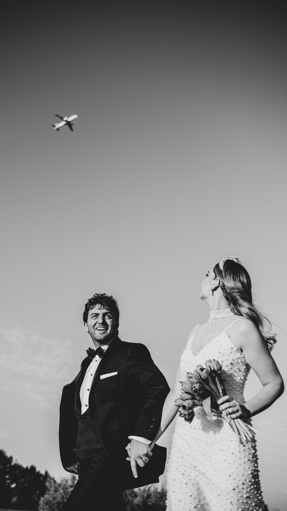 Black and white wedding photo of couple holding hands with airplane flying overhead.