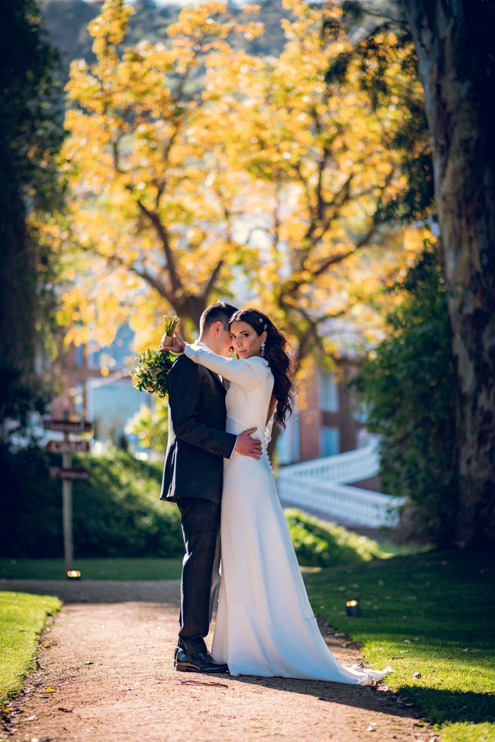 Bride and groom embrace on a garden path surrounded by vibrant autumn yellow trees.