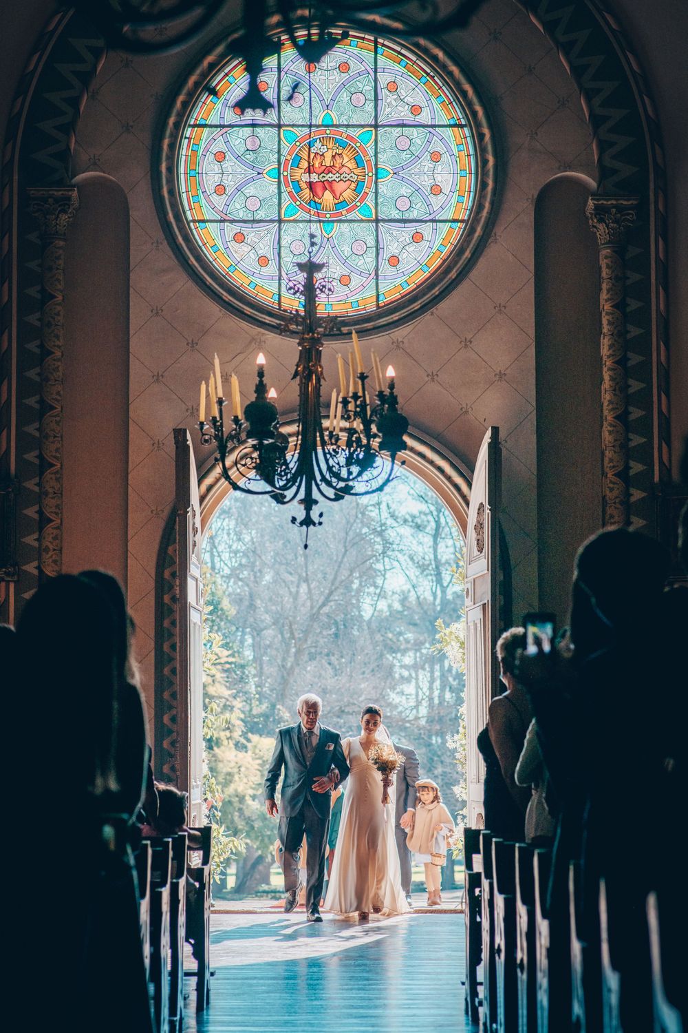 Bride walks down the aisle with escort beneath a stunning circular stained glass window in a chapel.