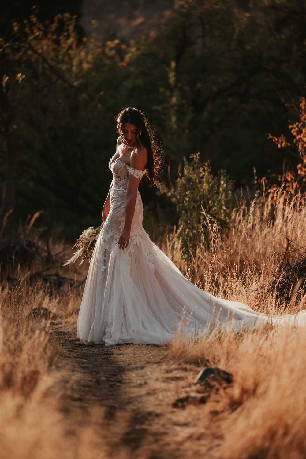 Bohemian bride in off-shoulder lace gown stands in golden dry grass field at sunset.