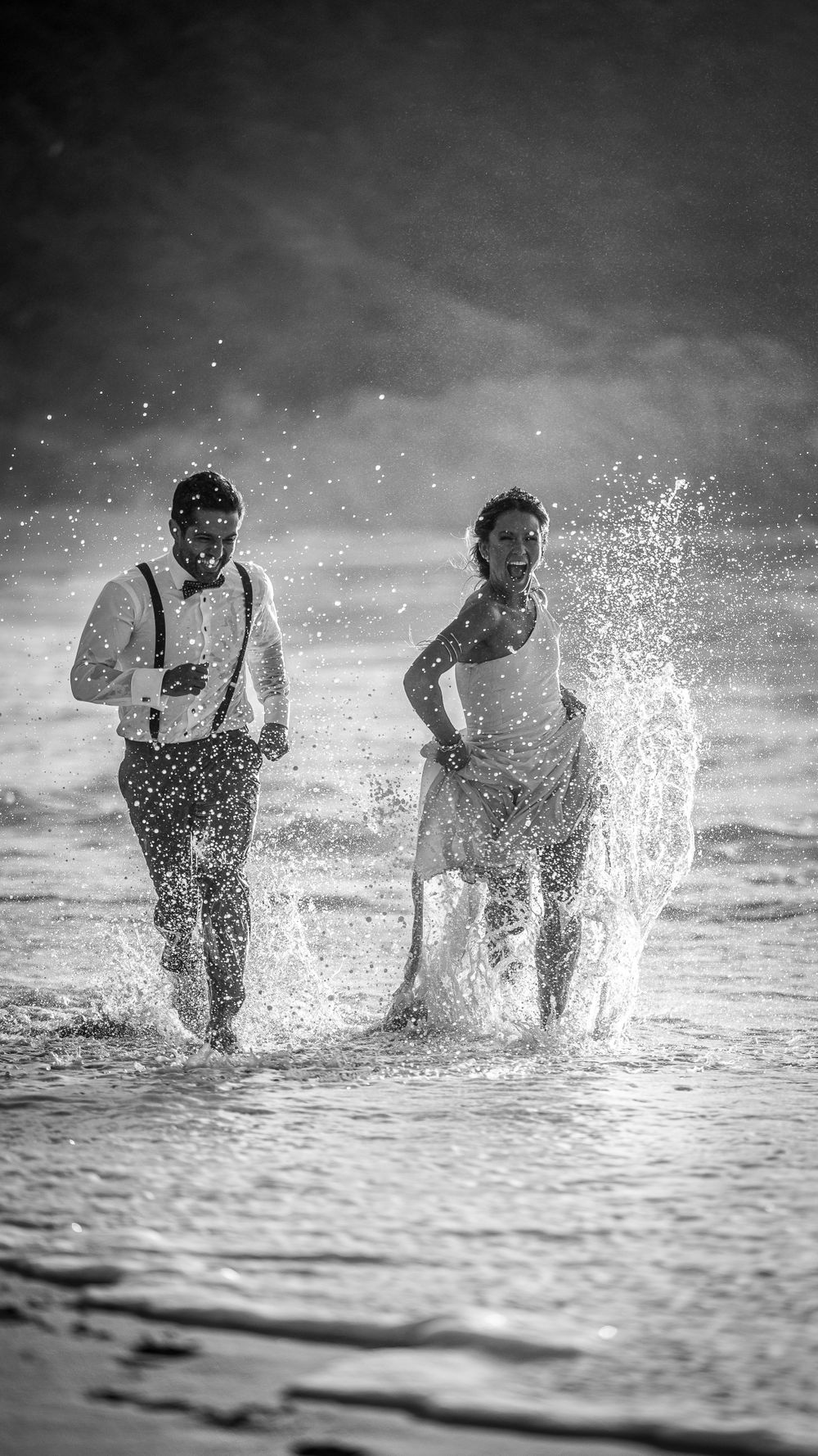 Black and white photo of bride and groom running joyfully through ocean waves on beach.