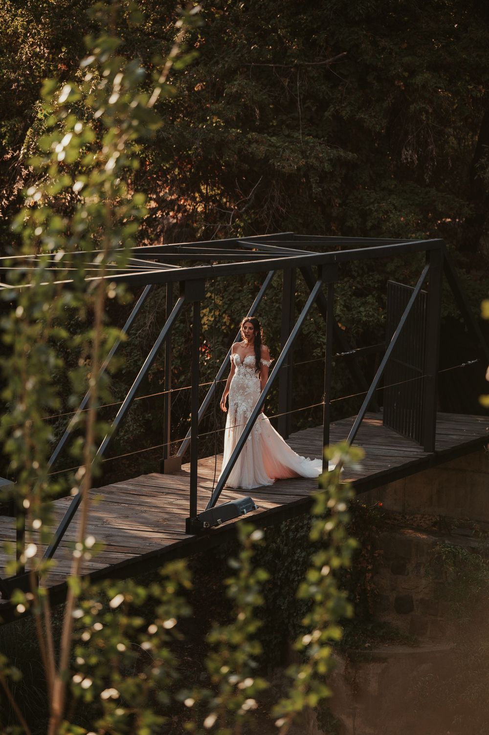 Bride in elegant white gown standing on a wooden bridge surrounded by lush green trees in golden light.