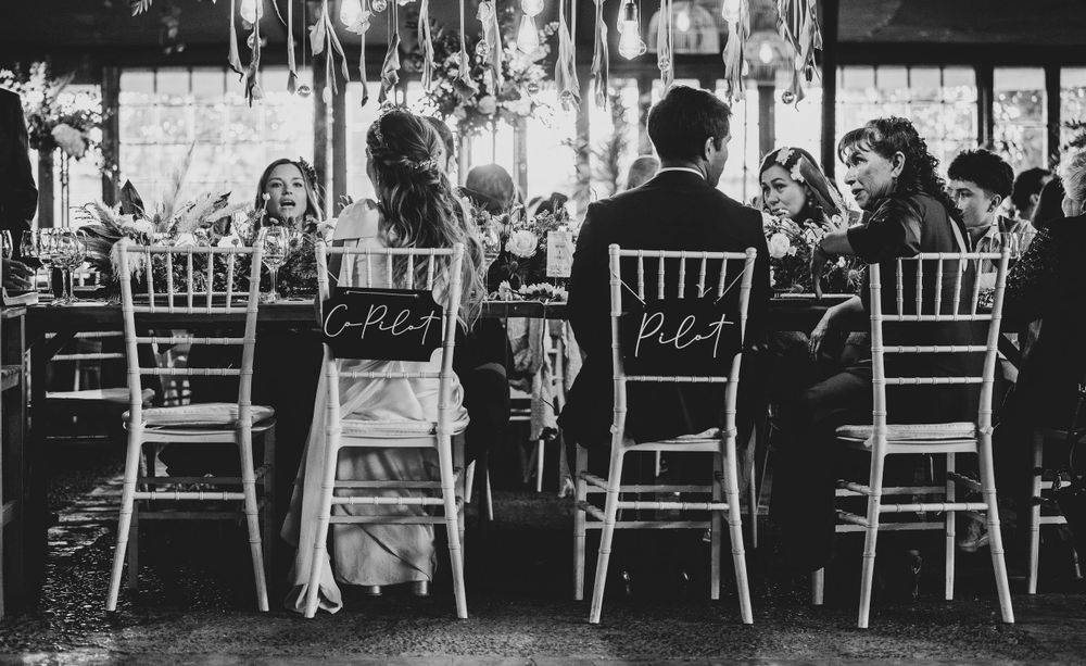 Black and white photo of wedding party seated at decorated reception table with personalized chair signs.