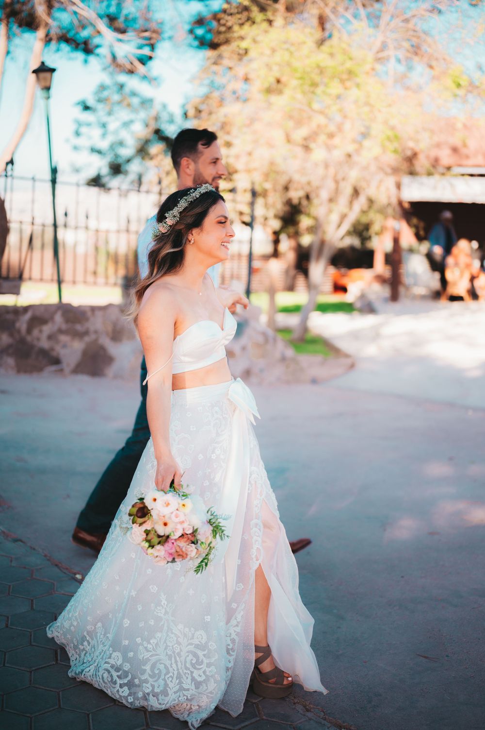 Smiling bride in lace two-piece wedding dress holding bouquet walks with groom in sunny outdoor setting.
