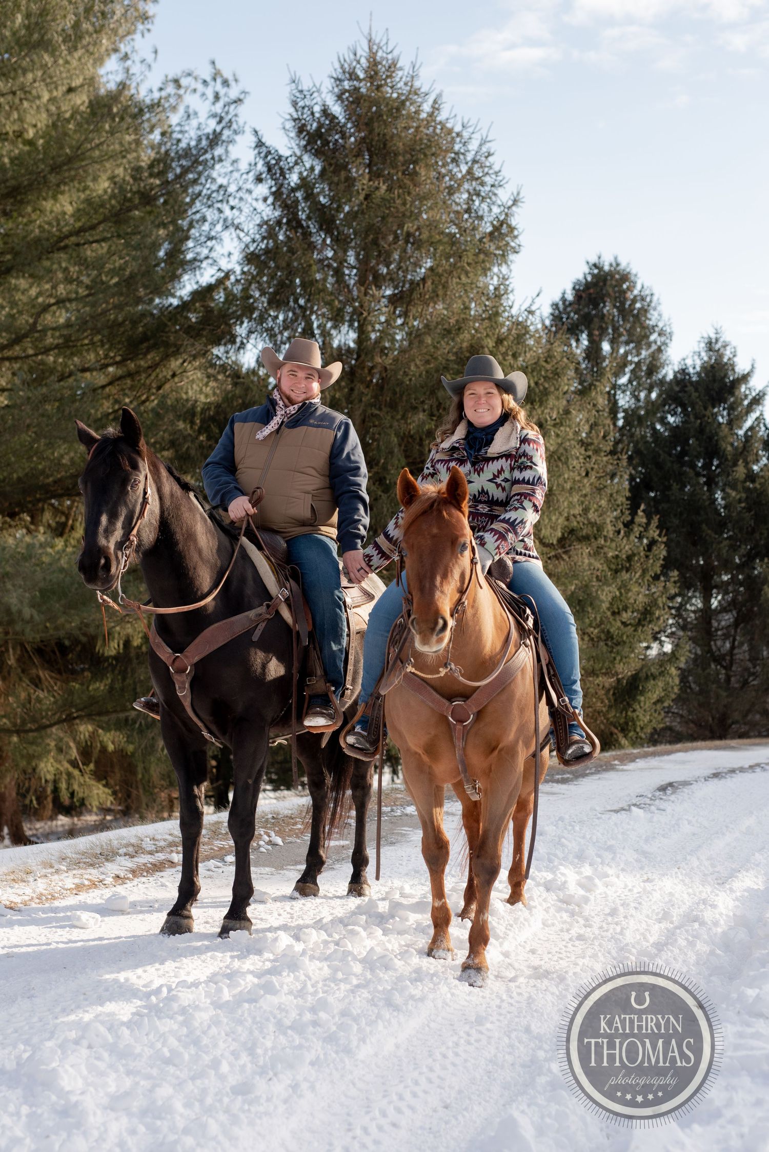 Two riders on horseback, one on a black horse and one on a brown horse, travel a snowy winter path.