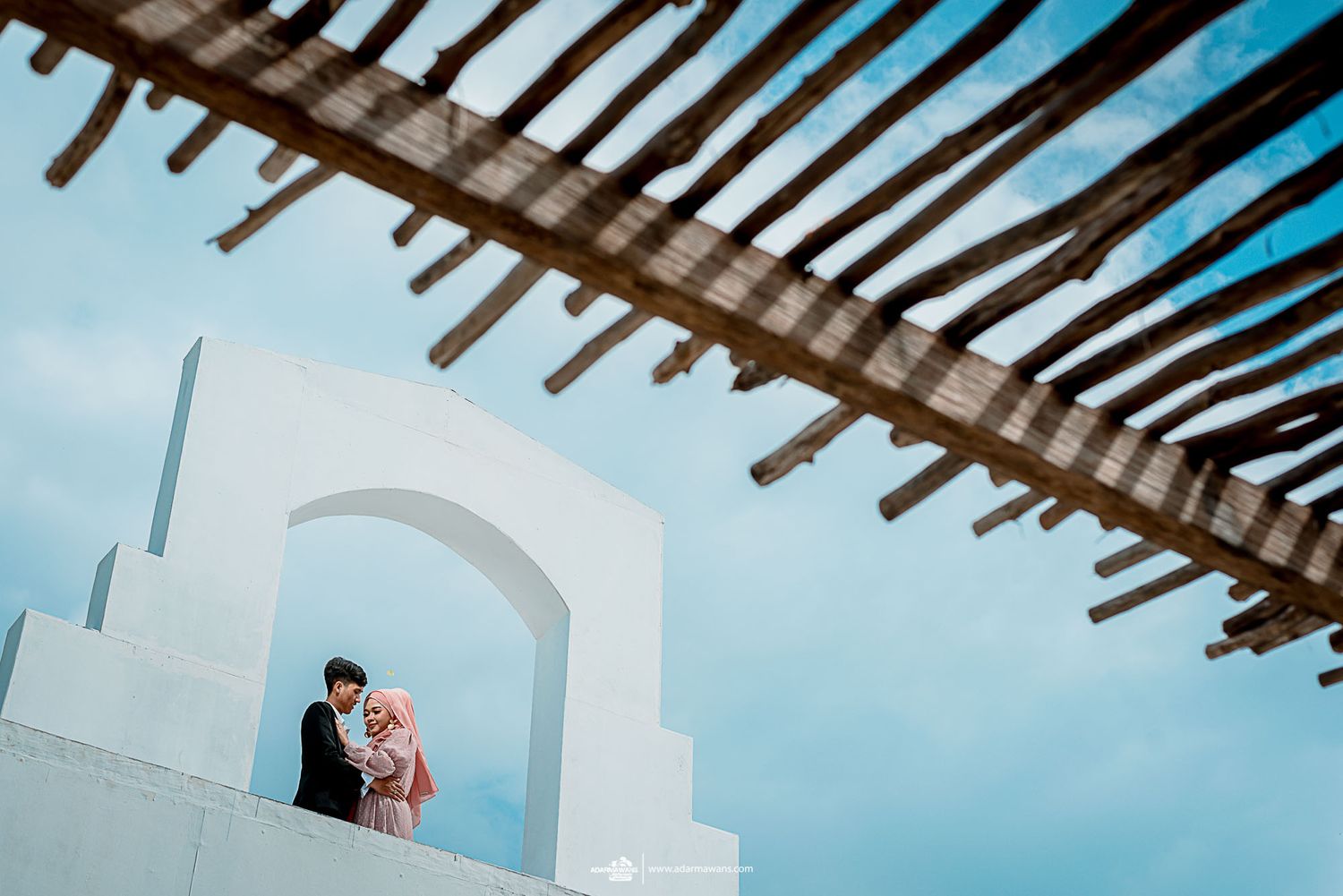 A romantic couple poses under a wooden pergola against a white archway and blue sky.