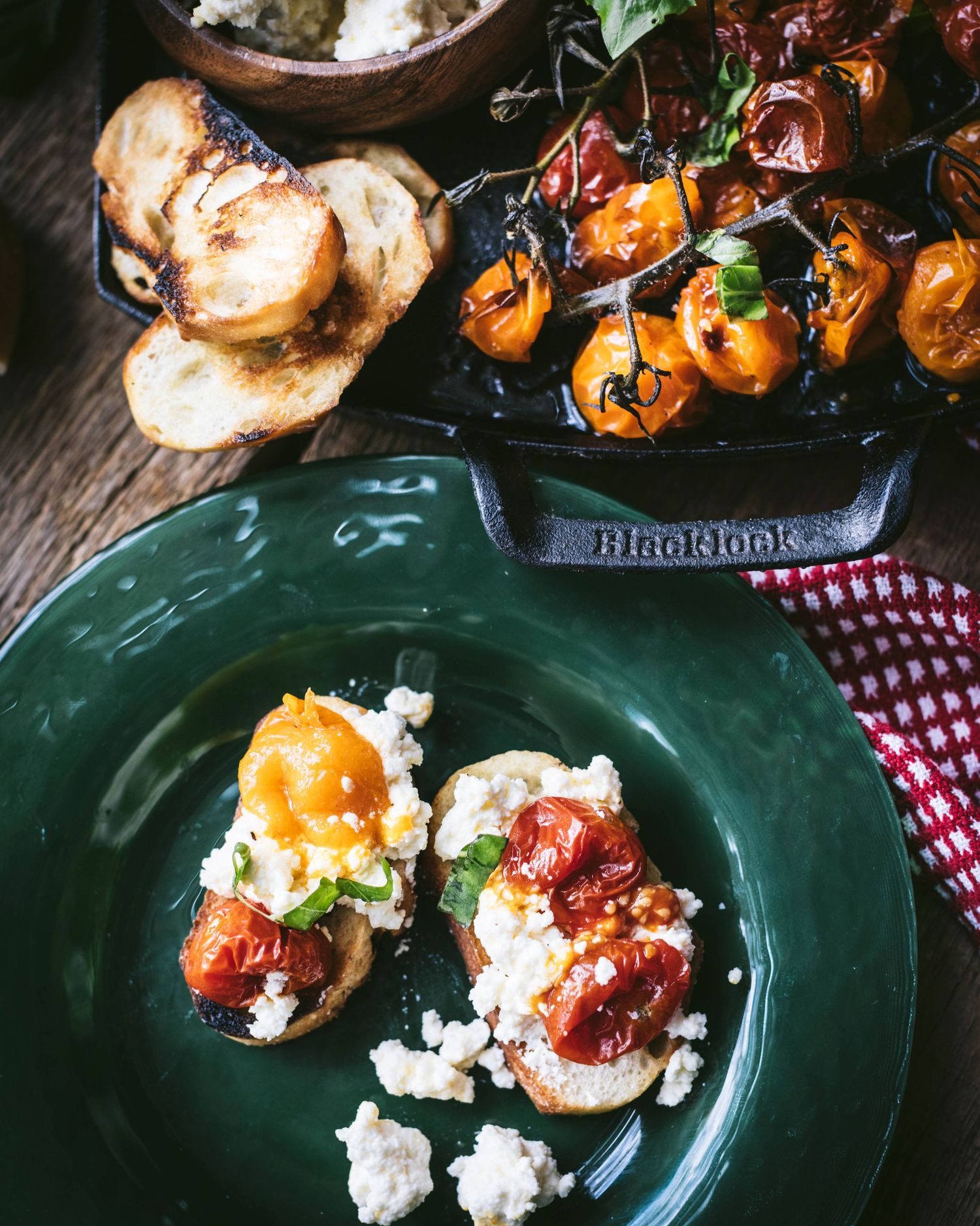 Bruschetta topped with creamy ricotta and roasted cherry tomatoes on a green plate with cast iron skillet behind.