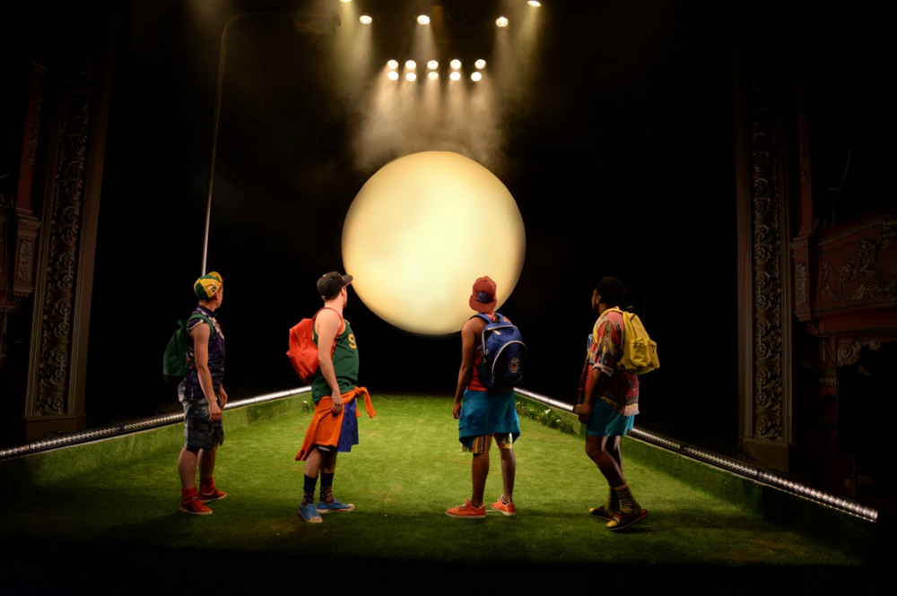 Performers in colorful costumes stand beneath stage lights and a glowing orb.