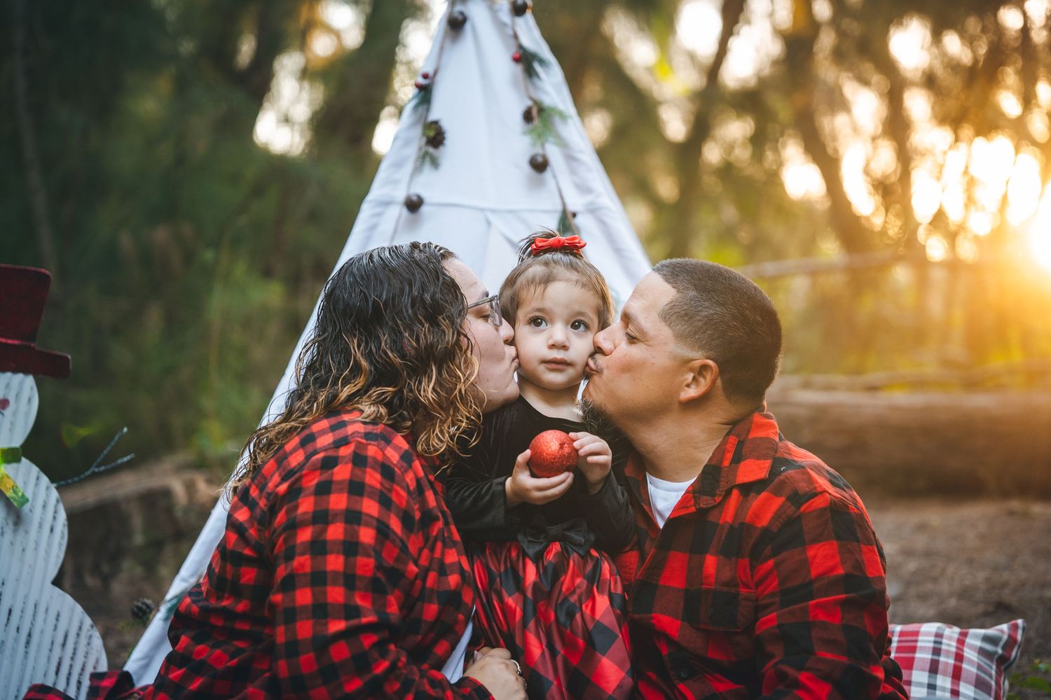 Family in matching red buffalo plaid flannel clothing sharing tender moment at sunset by teepee tent.