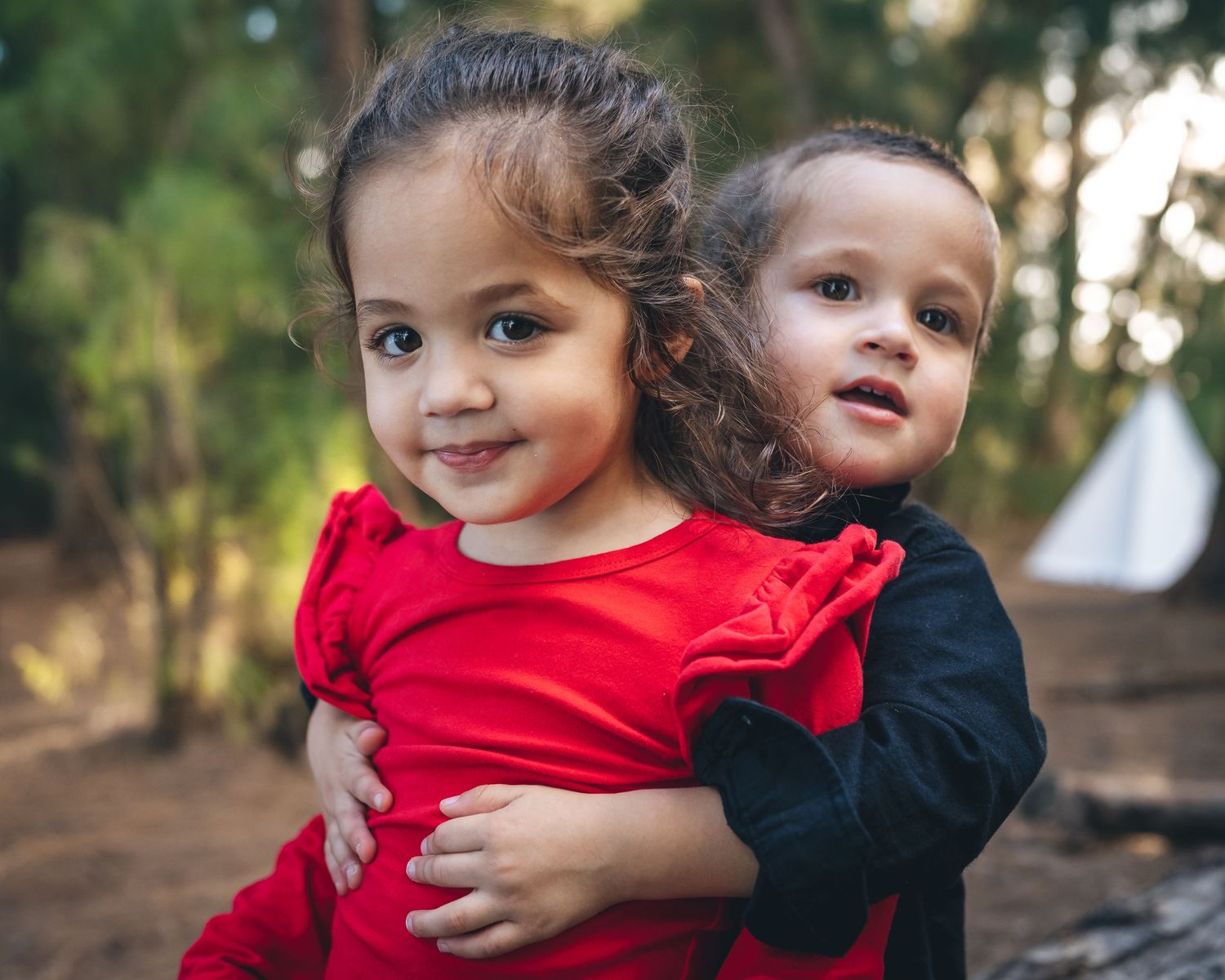 Two children in festive red and black outfits share sweet embrace outdoors.