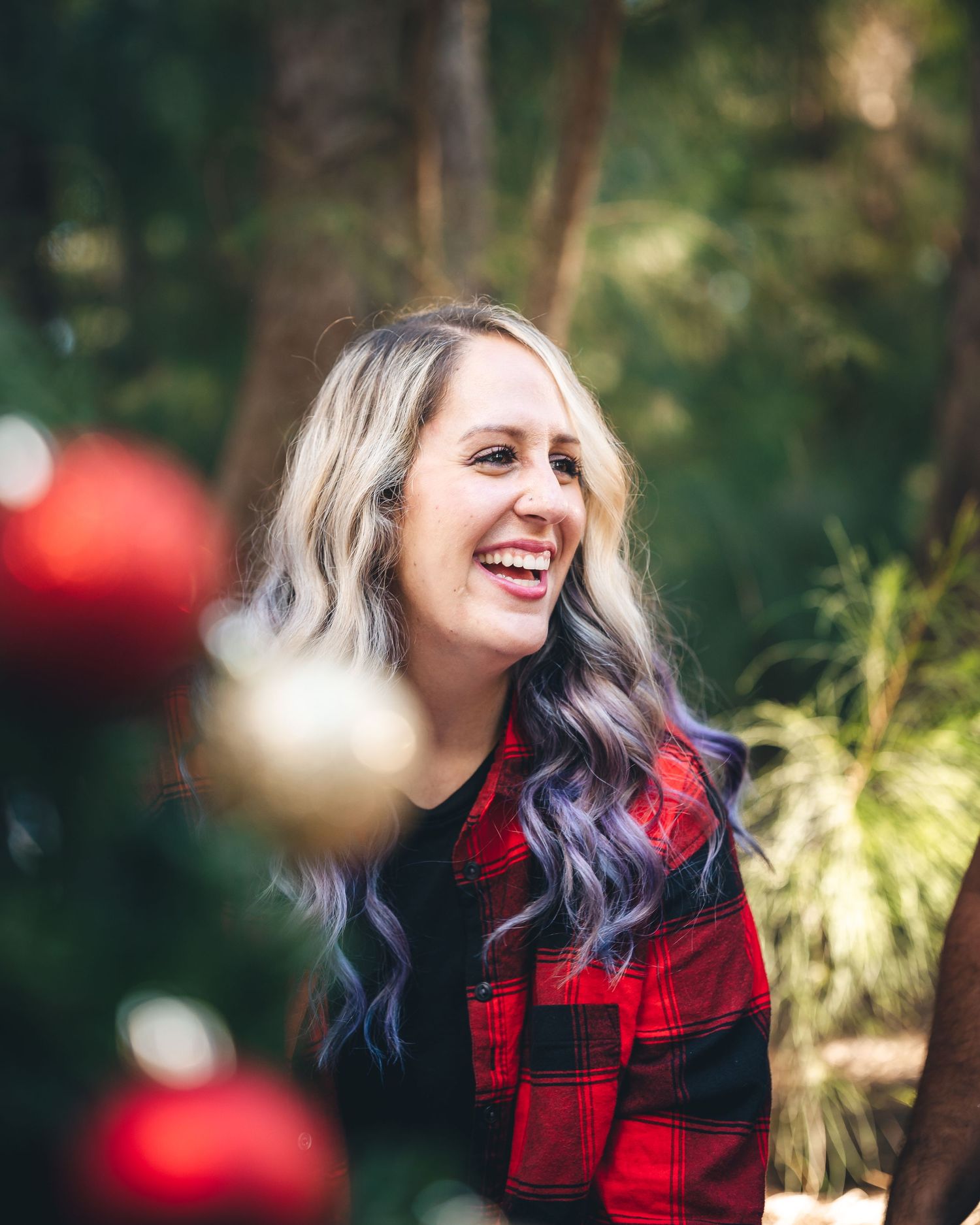 Woman in red plaid flannel laughs joyfully in forest setting with Christmas ornaments.