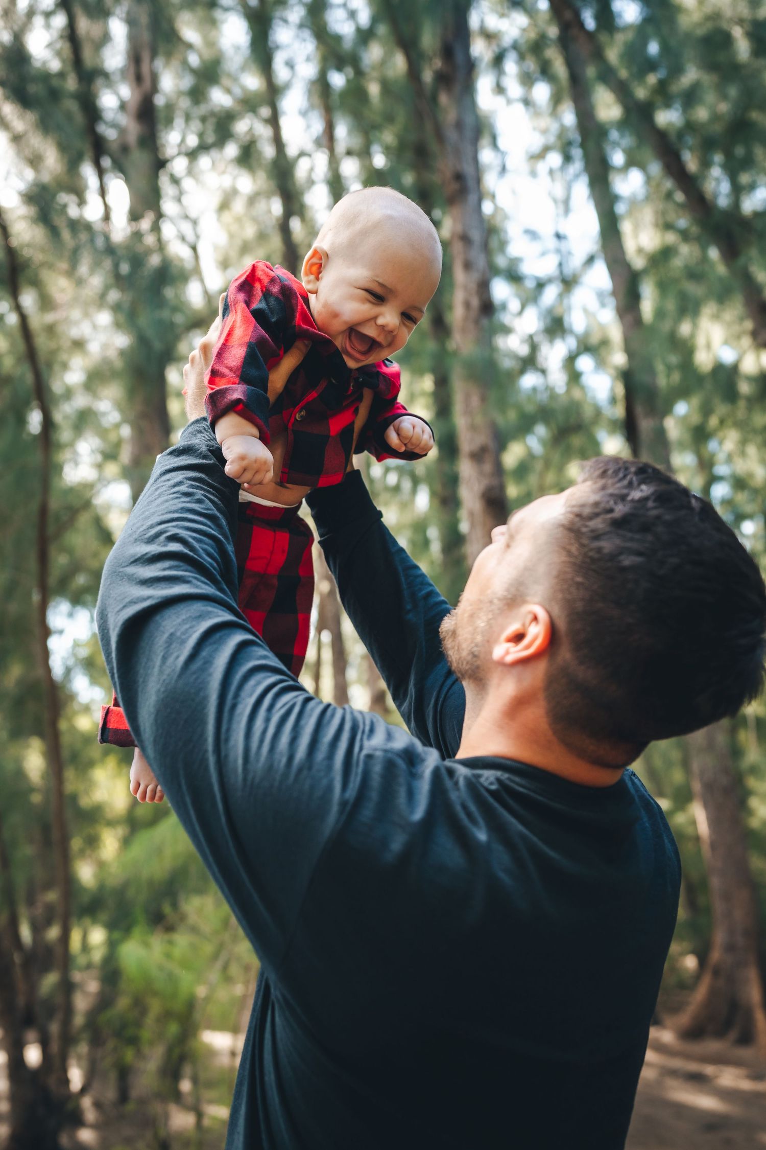 Parent lifts baby wearing red plaid outfit in forest setting.