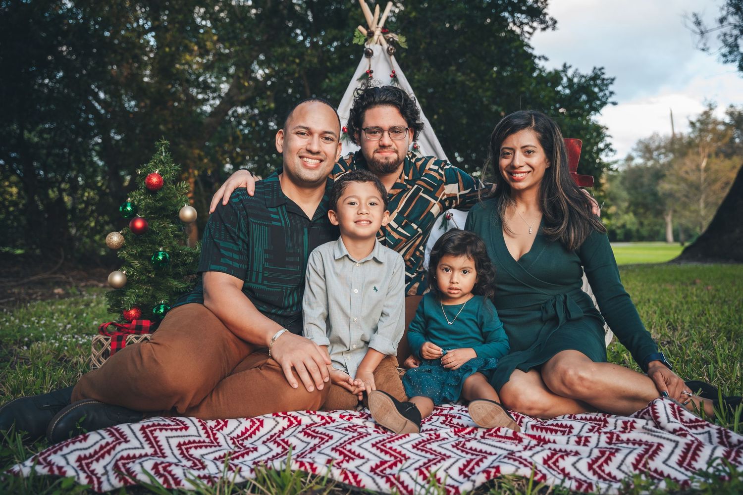 Family in coordinated outfits sitting on plaid blanket outdoors with decorated Christmas tree.