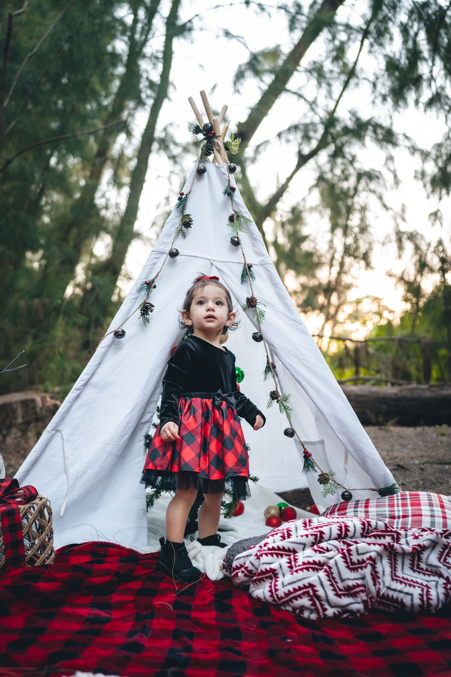 Small toddler in red and black outfit stands in front of white teepee decorated with string lights.