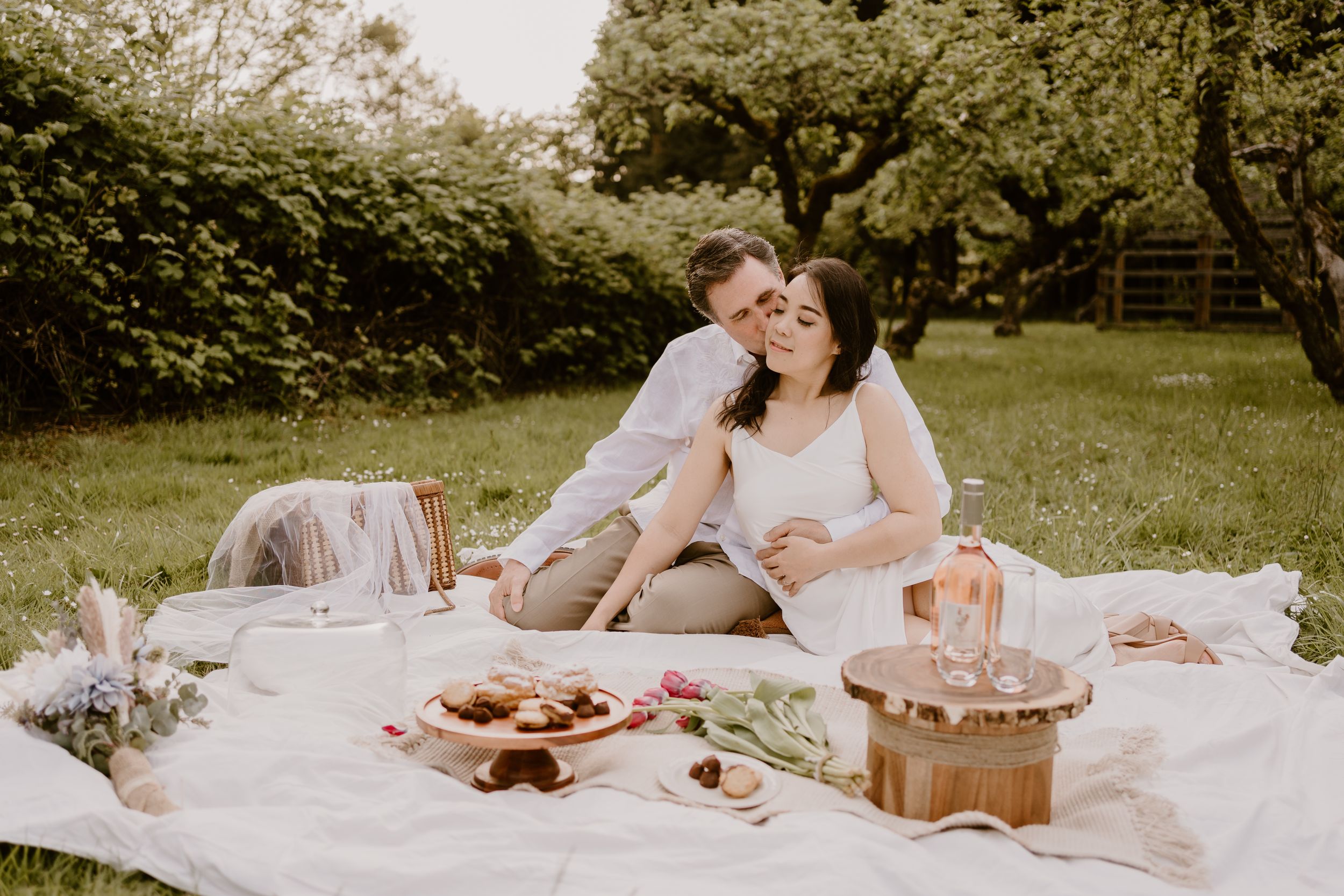 A romantic picnic setup on a white blanket in a grassy field with pastries and treats.