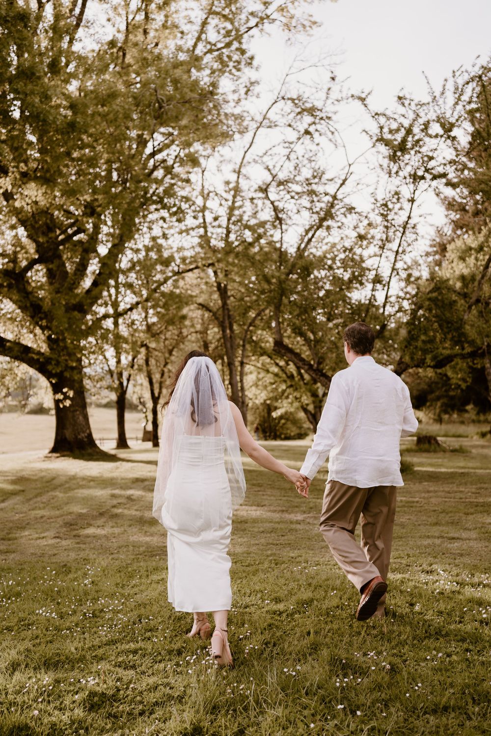 Two people in white outfits walking hand in hand through a grassy field at sunset.