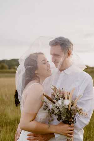 A couple shares an intimate moment in a field while holding a floral bouquet.