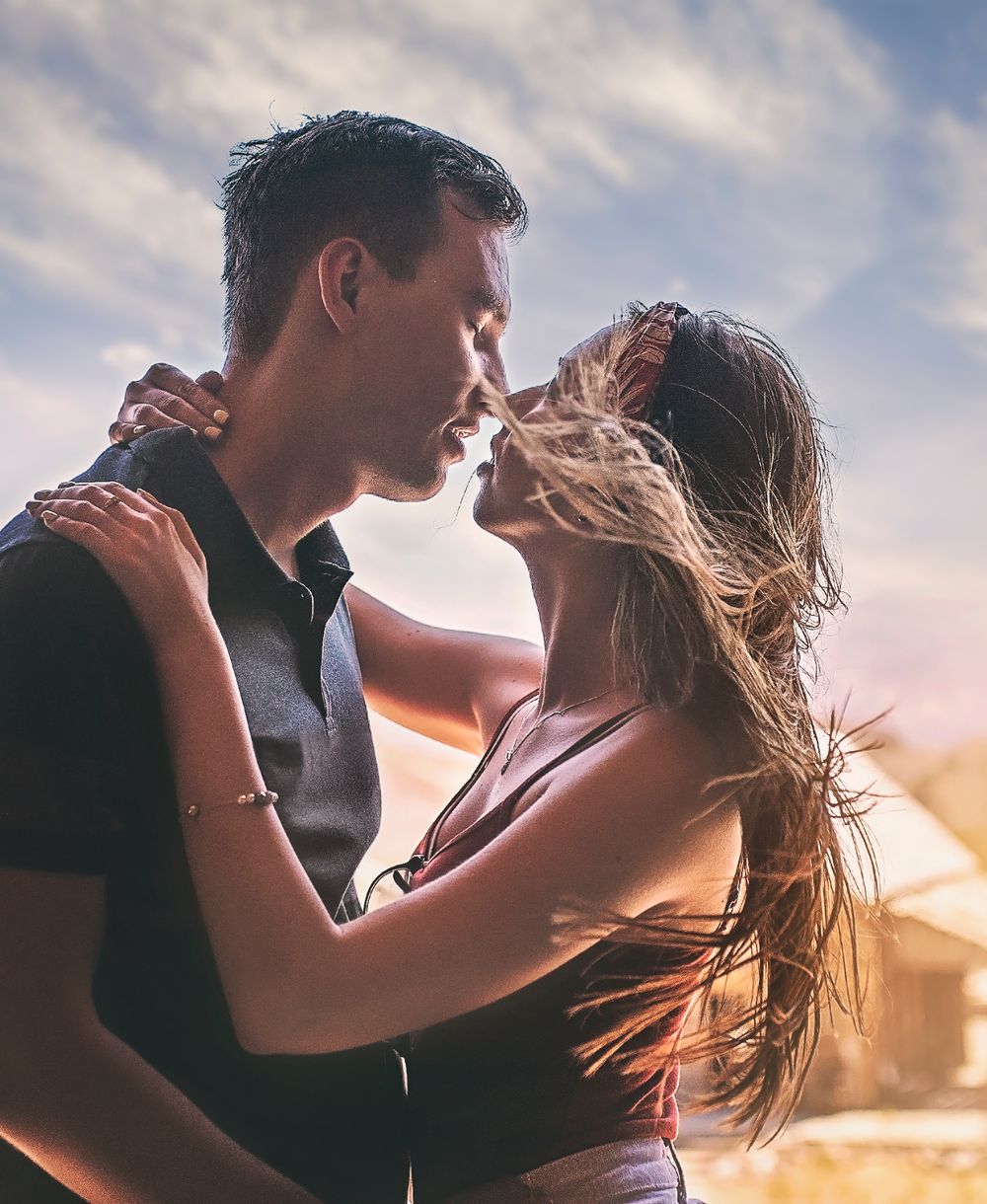 A romantic silhouetted couple embraces against a sunset sky with wind-blown hair.