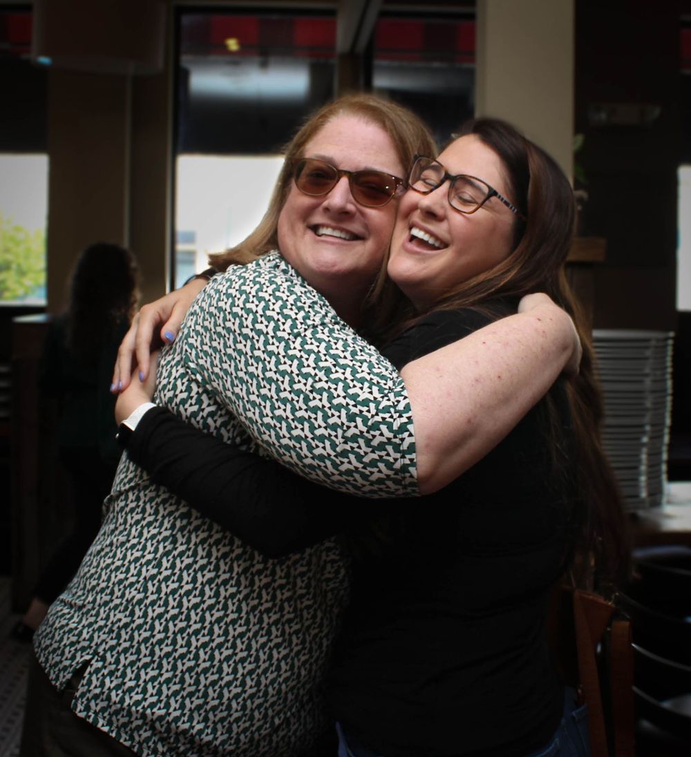 Two people share a warm embrace and laugh together at an indoor gathering.