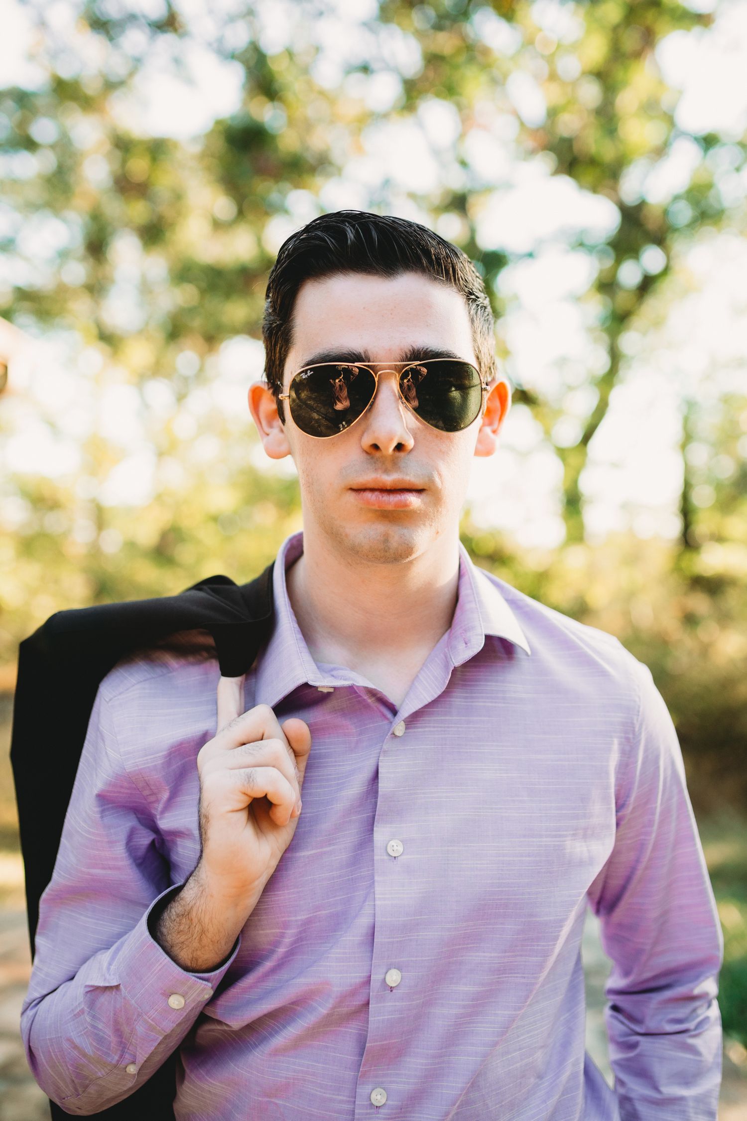 a high school senior stares directly at the camera; he wears sunglasses, a purple shirt, and holds a suit in one hand