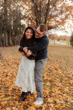 A couple embraces while standing among fallen autumn leaves in a wooded area.