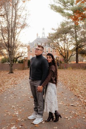 A couple poses in front of a historic colonial building on an autumn day with fallen leaves.