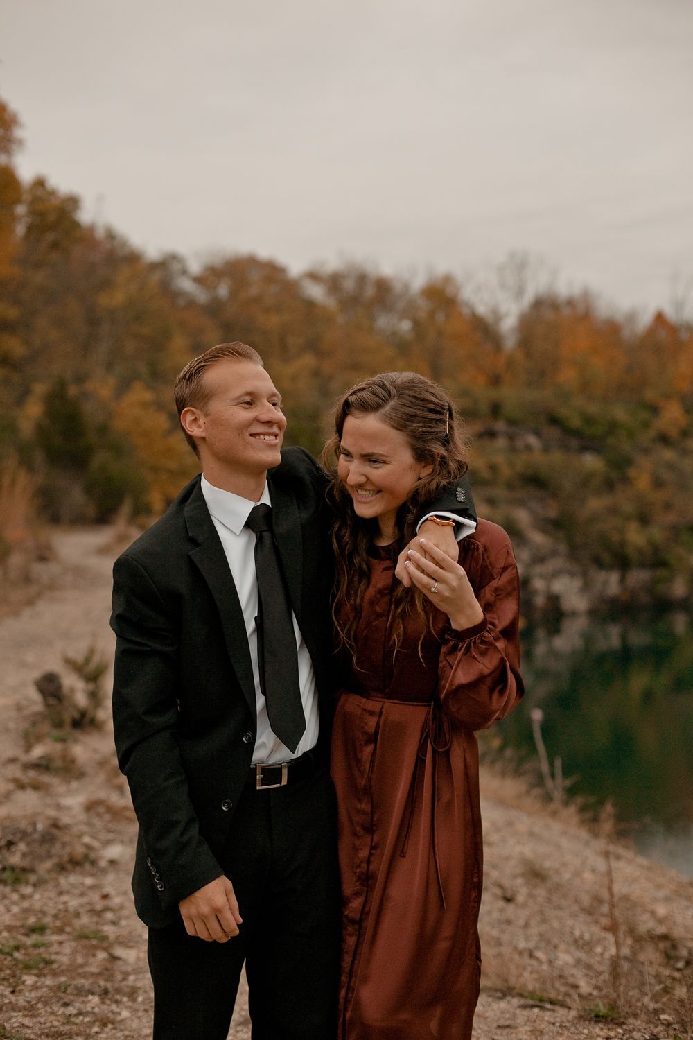 A couple laughs together while standing near a scenic lake with fall colors in the background.