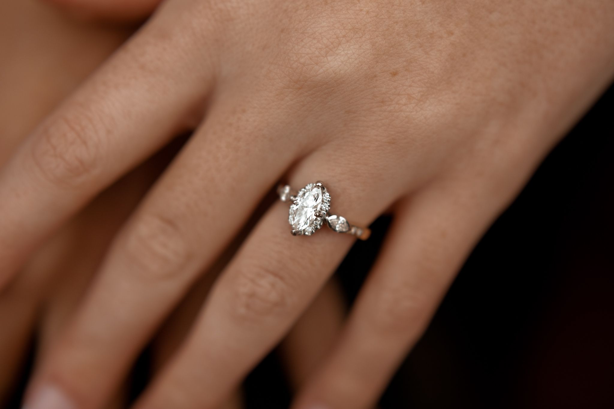 Close-up details of an engagement ring on a hand against a dark background.