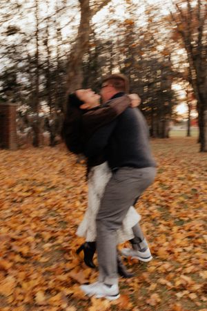 A couple shares a joyful embrace in a park covered with fallen autumn leaves.