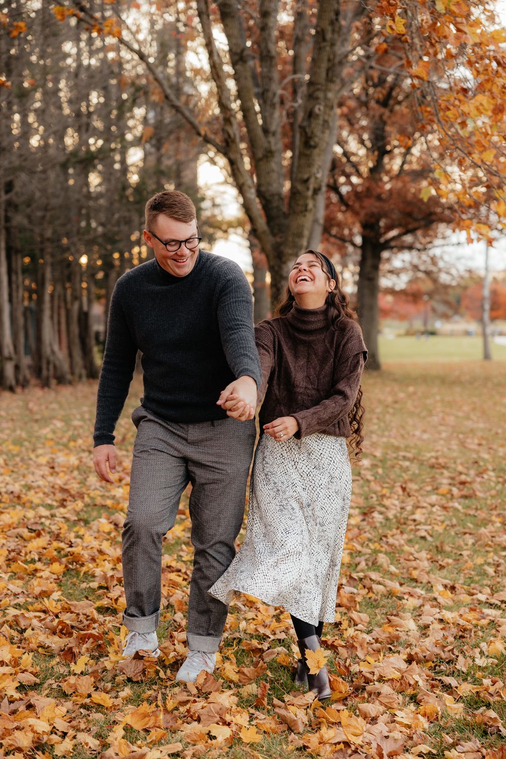 A couple playfully walks through fallen autumn leaves in a park setting.