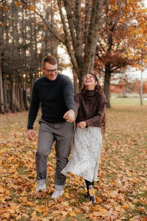 A couple playfully walks through fallen autumn leaves in a park setting.