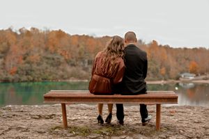 Two people sit together on a wooden bench overlooking an autumn lake.