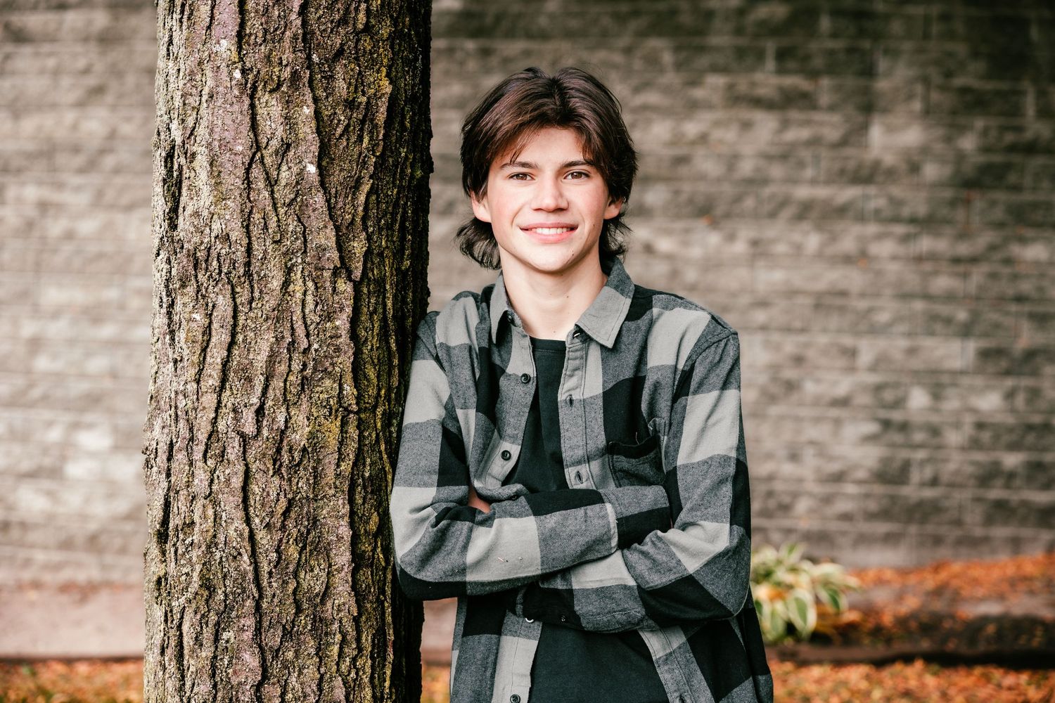 Portrait of person in plaid flannel shirt leaning against tree trunk with brick wall background.