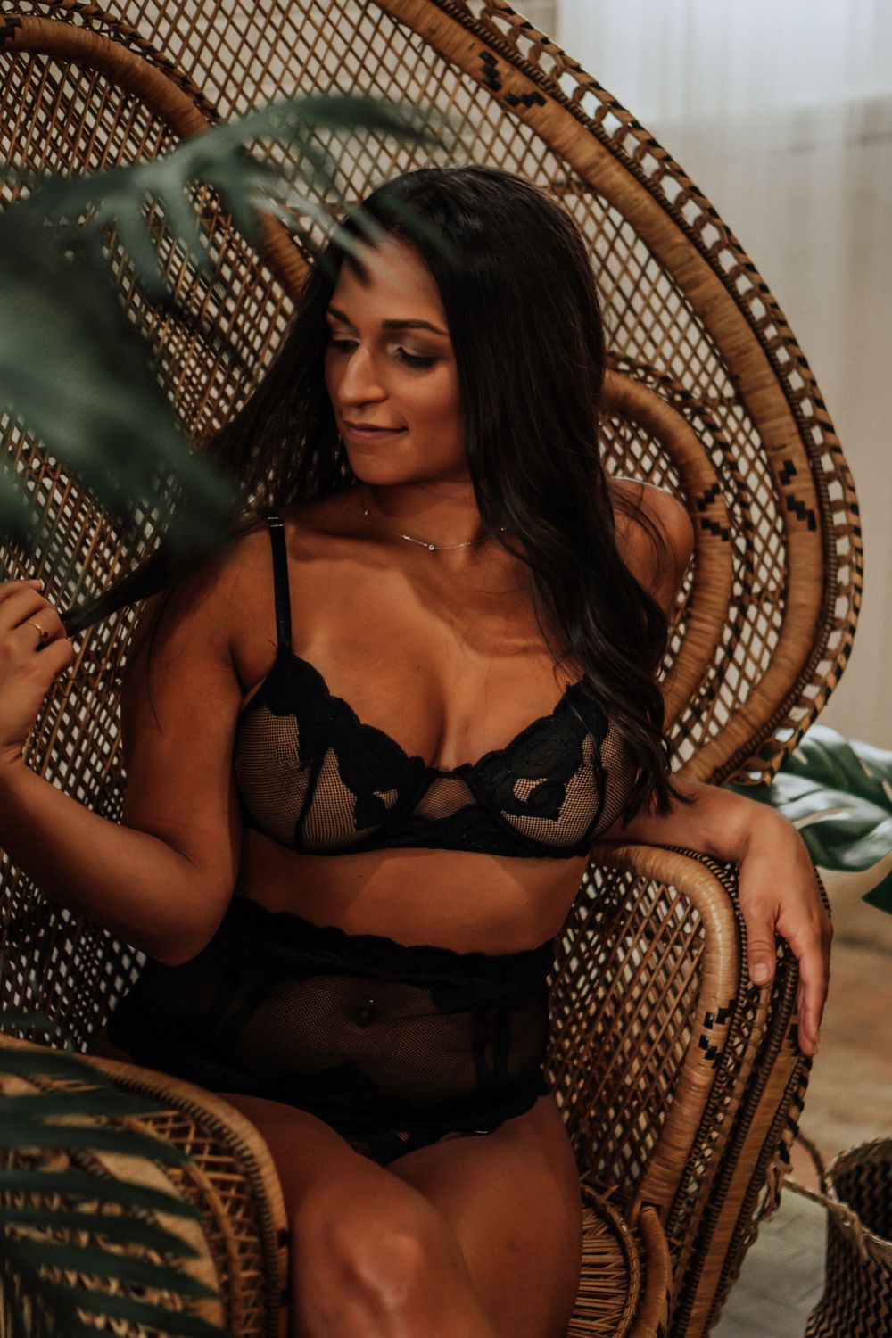 Woman in black lace lingerie seated in wicker peacock chair surrounded by tropical leaves.