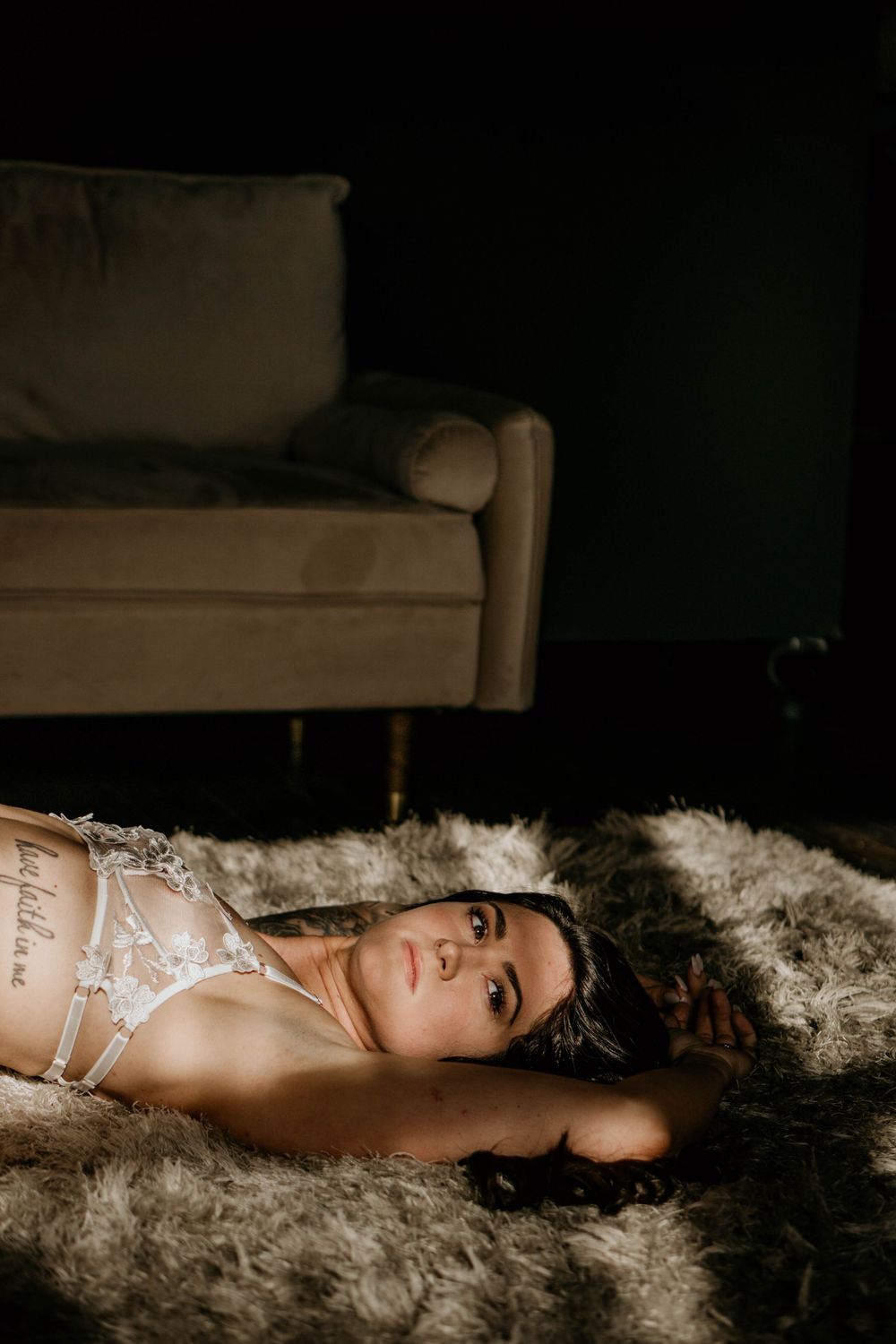Woman in white lingerie lying on a fur rug in moody lighting with a couch in the background.