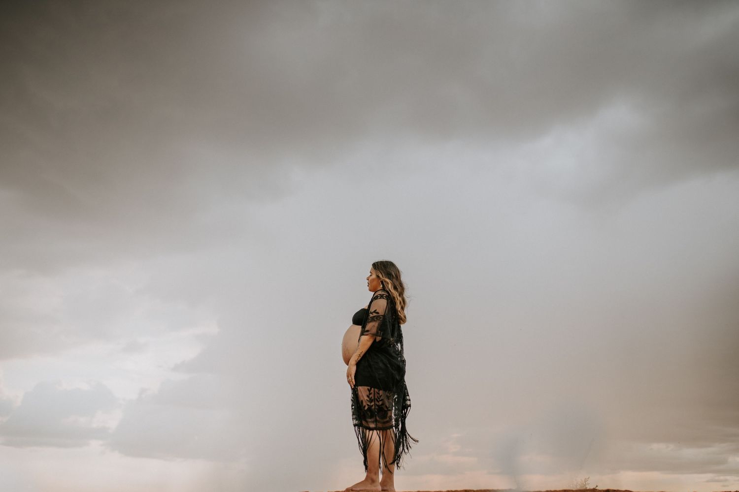 Beautiful mama to be poses on a hill at Granite Mines, Mount Isa as the rain falls behind her looking down at her bump