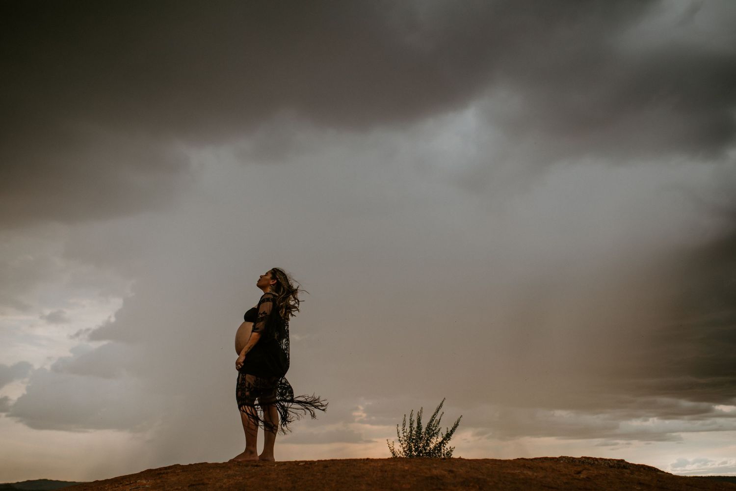 Beautiful mama to be poses on a hill at Granite Mines, Mount Isa as the rain falls behind her