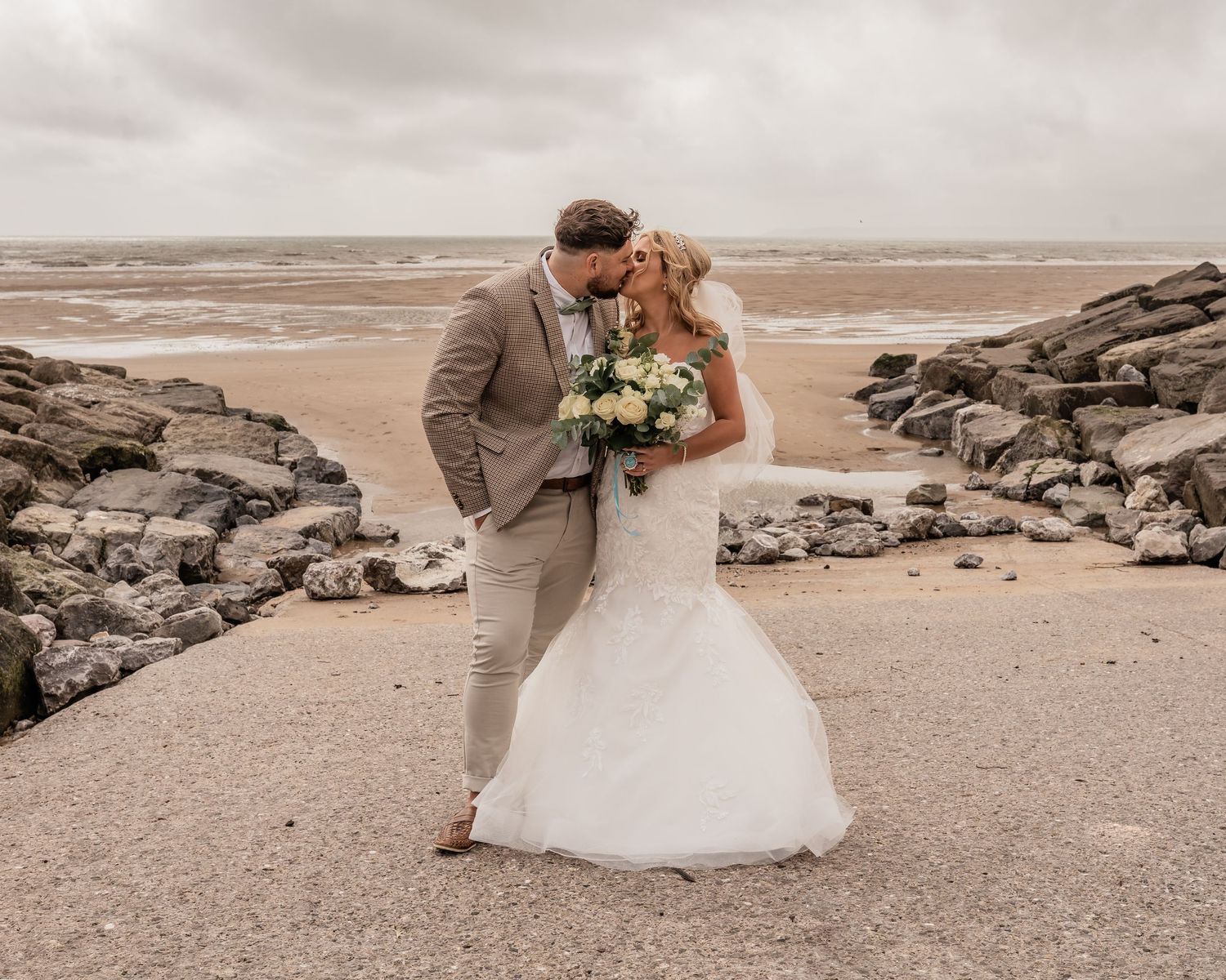 Newlyweds share an intimate moment on a rocky beach while holding a bridal bouquet.