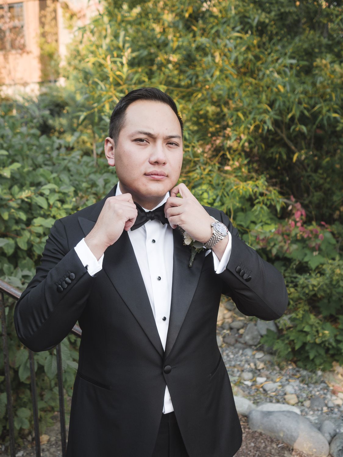 Groom adjusts a black bow tie while wearing a formal black tuxedo with white shirt in an outdoor garden setting.