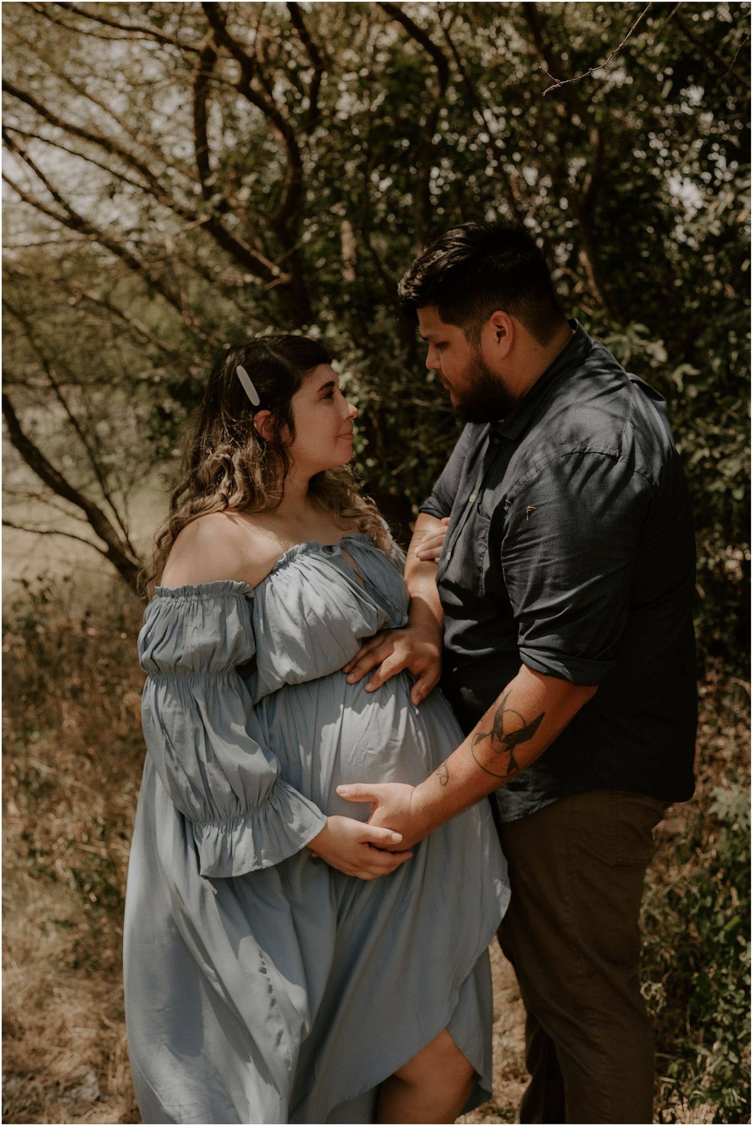 Candid maternity moment captured between couple wearing blue dress and dark shirt outdoors.