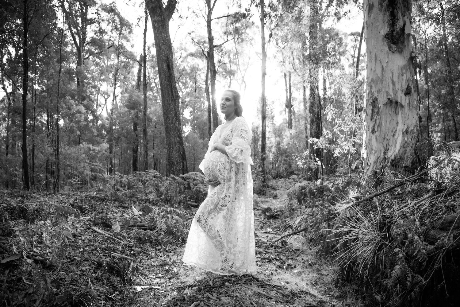 Black and white maternity portrait of a woman in a flowing dress standing among tall trees.