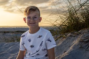 Young boy in a t-shirt with dinosaur prints sits on sandy dunes at golden hour.