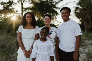 Four people in white outfits pose together outdoors during golden hour with sunlight filtering through trees.