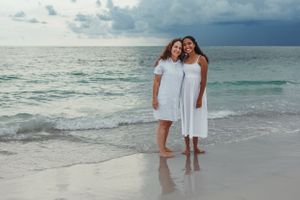 Two people in white dresses embrace while standing on a tropical beach at sunset.